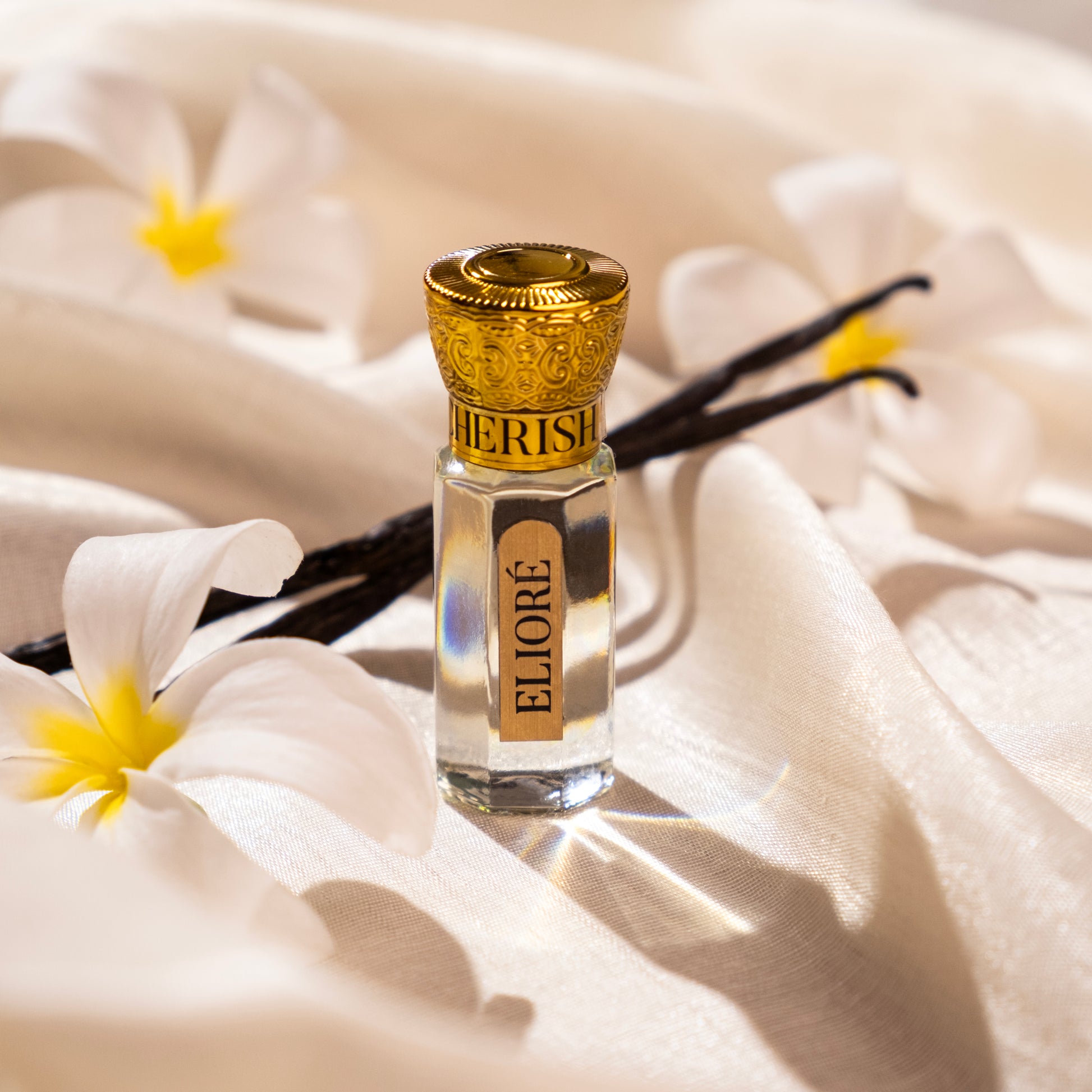 Cherish by Elioré luxury attar perfume bottle styled with vanilla pods and white flowers on soft silk fabric for an elegant gourmand fragrance aesthetic.