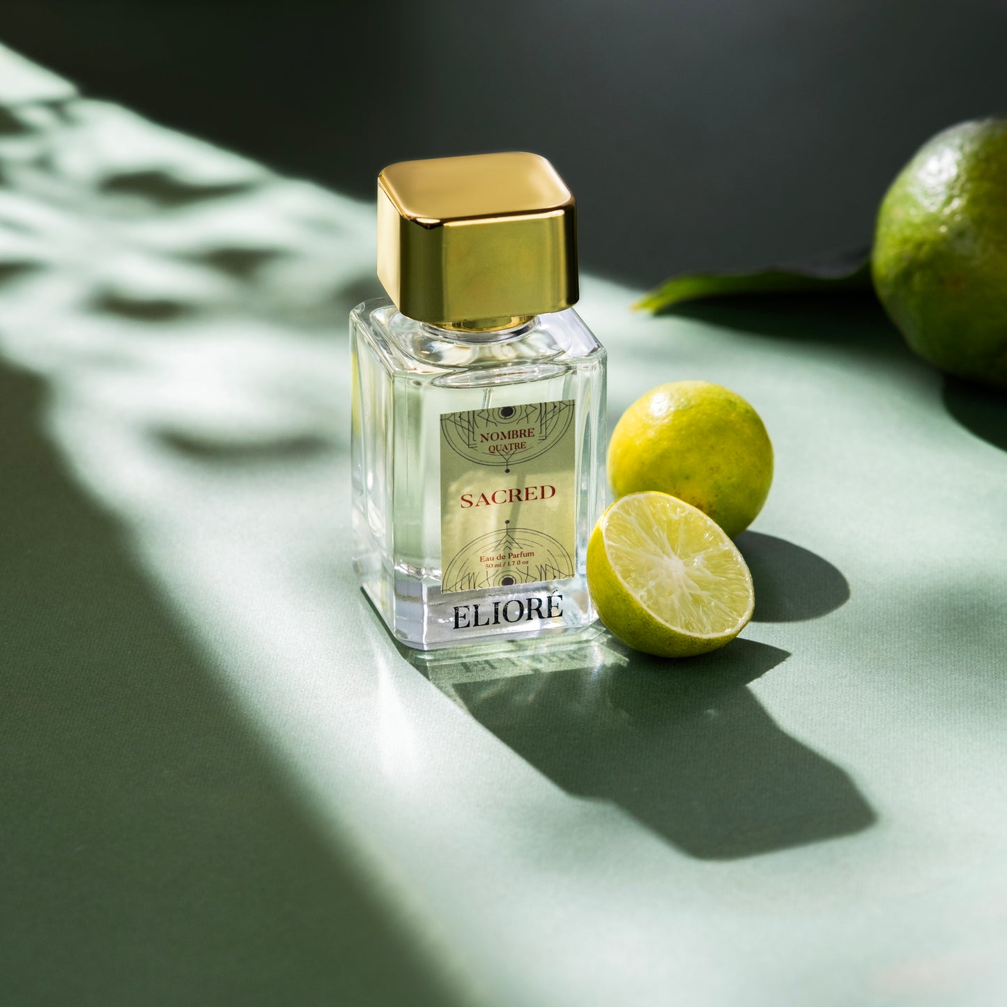 Perfume bottle labeled 'Eliore' with gold cap and 'Sacred' text, surrounded by limes on a green surface.