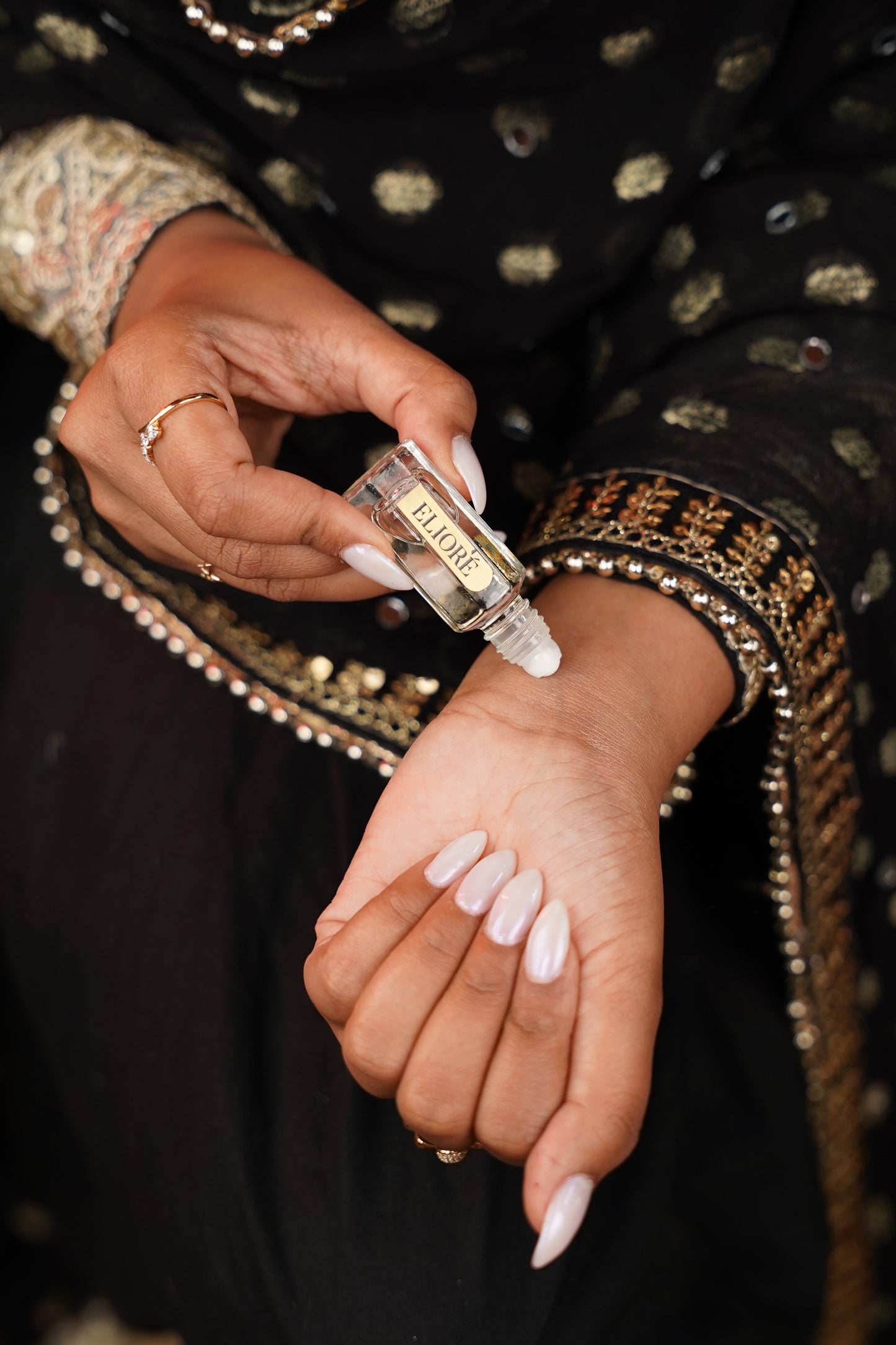 Person applying a clear liquid to their wrist with a small bottle, wearing a black and gold patterned garment.