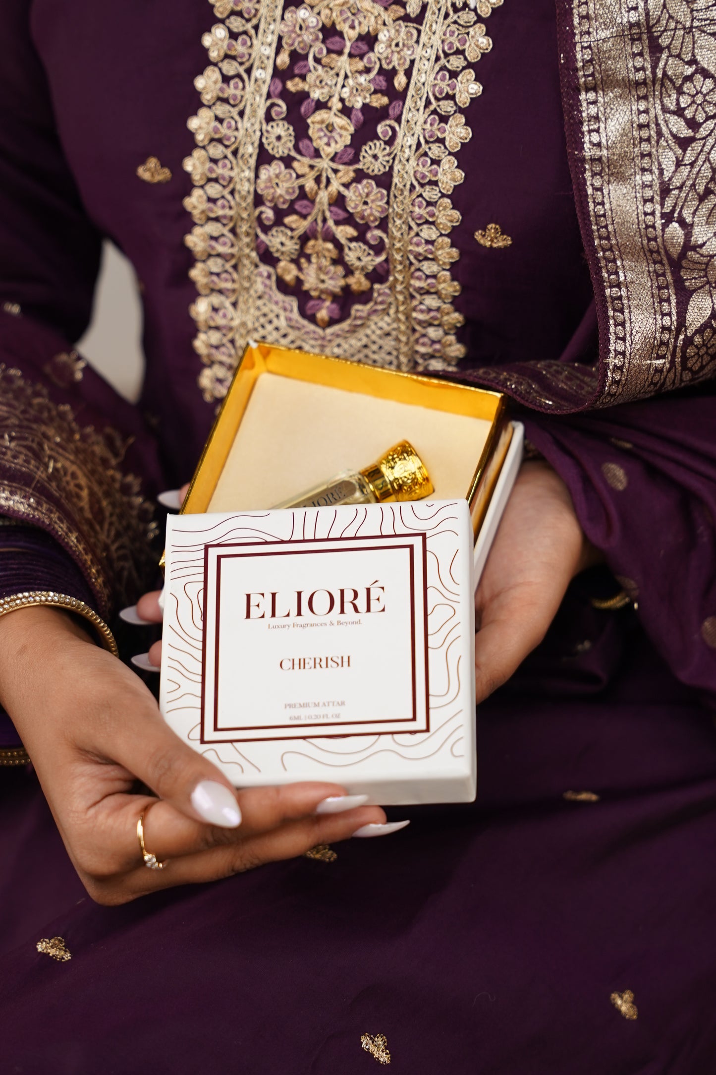 Hand holding a box of Eliore cherish attar with intricate purple fabric background