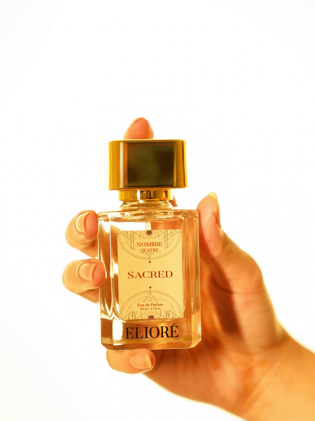 Hand holding a gold and clear sacred perfume bottle labeled 'Eliore' on a white background